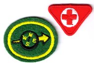 ovales Leistungsabzeichen, Grundfarbe grün, gelber Kreis ringsrum, im Inneren ein kleiner Kompass, symbolisiert das Abzeichen Orientierung. Ein kleines rotes dreieckiges Abzeichen, mit weißem Kreis und darin einem roten Kreuz steht für Erste Hilfe der Jungpfadfinder